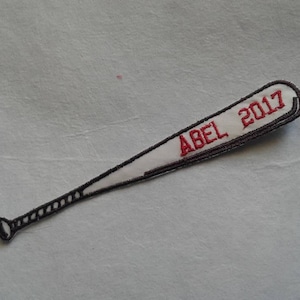May include: A white embroidered baseball bat with black stitching and the text "ABEL 2017" in red thread.