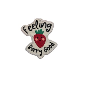Può includere: Toppa ricamata con bordo bianco e le parole "Feeling Berry Good". La toppa presenta una fragola rossa sorridente con foglie verdi. Il testo e la fragola sono centrati sulla toppa.