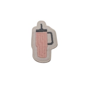 Könnte beinhalten: Ein gestickter Aufnäher, der einen rosa Becher mit Strohhalm in den Farben Rosa und Grau zeigt.