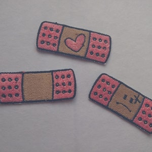 May include: Three embroidered iron-on patches shaped like band-aids. One has a heart, one has a sad face, and one has a plus sign.