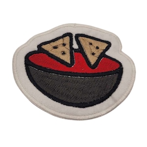 May include: A white embroidered patch with a black bowl filled with red salsa and two brown tortilla chips.