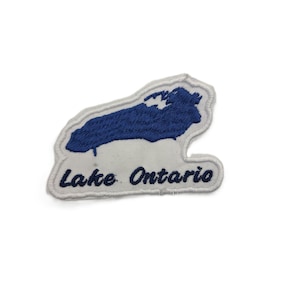 Op de afbeelding: Een witte geborduurde patch met een blauwe kaart van het Lake Ontario. De patch heeft een witte rand en de tekst Lake Ontario is in donkerblauw cursief schrift onder de kaartvorm op de witte stoffen achtergrond geborduurd.