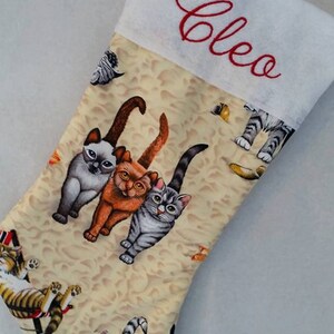 May include: A white Christmas stocking with a red embroidered name "Cleo" on the cuff. The stocking is decorated with a beige fabric featuring a repeating pattern of cats.