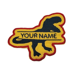 May include: Yellow and red embroidered iron-on patch featuring a black silhouette of a dinosaur with the text "YOUR NAME" in black.