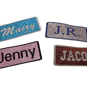 Könnte beinhalten: Vier rechteckige Glitzeraufnäher mit gestickten Namen: "Maizy" in Weiß auf einem blauen Aufnäher, "J.R." in Blau auf einem silbernen Aufnäher, "Jenny" in Schwarz auf einem rosa Aufnäher und "Jacob" in Weiß auf einem roten Aufnäher.