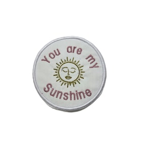 May include: A circular white patch embroidered with the words "You are my Sunshine" in pink. The design includes a gold sun with closed eyes and a smiling mouth. The patch has a white border.