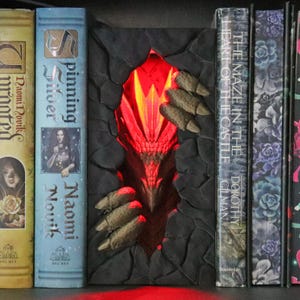 May include: A book nook insert featuring a dark, cracked stone wall texture with a glowing red dragon head emerging from the center. The dragon has textured scales and sharp claws gripping the edges of the stone opening, illuminated by a bright red light.