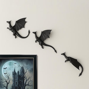May include: Three matte black dragon wall sculptures in varying flight poses, each featuring detailed wings, textured scales, and long tails. Below the dragons is a framed rectangular print depicting a dark castle silhouette under a full moon.