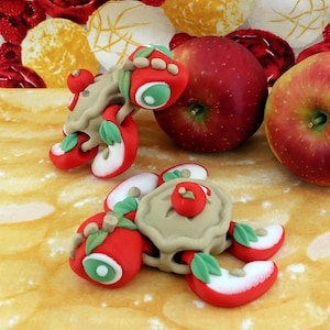 May include: Two small turtle figurines crafted from polymer clay, featuring shells shaped like miniature apple pies topped with red apples. The turtle heads and limbs are decorated with red apple slices and green leaves, positioned near two whole red apples.