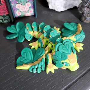 May include: A flexible articulated lizard figurine featuring a bright lime green body with tan accents and dark forest green scales shaped like clover leaves along its back. The creature has a smooth head with white eyes and three-toed feet.