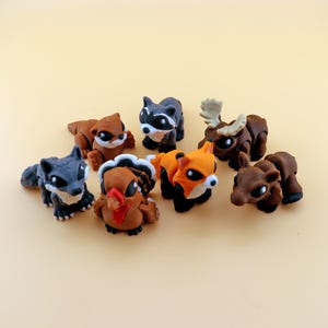 May include: A collection of small, colorful animal figurines, including a wolf, raccoon, fox, turkey, moose, bear, and otter. The figurines are in various shades of brown, orange, gray, and black, with white accents. They are arranged on a light yellow background.