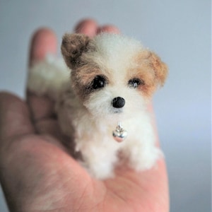 May include: A small, handmade white and brown dog figurine with a silver bell collar. The miniature dog is held in a person's hand, showcasing its fluffy texture and detailed features. The dog's ears and patches are brown.
