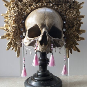 May include: A gold skull with a decorative crown and pink tassels hanging from a chain. The skull is on a black stand.