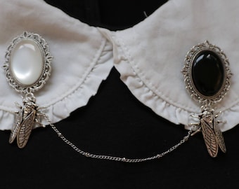 May include: Two silver brooches with a chain connecting them. One brooch has a white oval cabochon and the other has a black oval cabochon. Both brooches feature a detailed design and a cicada pendant.