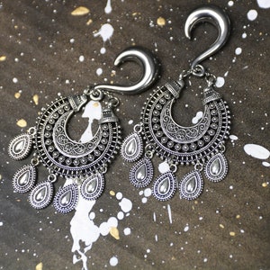 Könnte beinhalten: Ein Paar silberne Ohrdehner mit verzierten, baumelnden Anhängern. Die Dehnungen sind aus Metall und haben ein Haken-Design. Die Anhänger sind tropfenförmig und haben ein Perlen-Design.