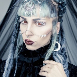 May include: A woman with blue hair and silver jewelry, wearing a black lace top and a silver crescent moon earring. She has a silver nose ring and a silver chain across her forehead.
