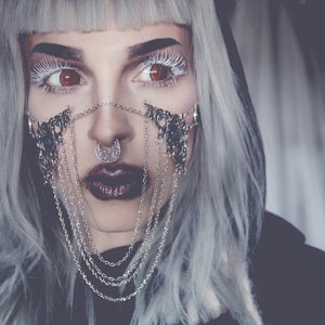 May include: A woman with silver hair and dark makeup wears a silver chain nose ring and a silver chain face mask. The mask is made of delicate chains and small metal embellishments.