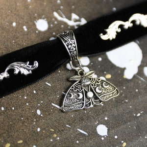 May include: A black velvet choker necklace with a silver pendant featuring a detailed moth design with crescent moons. The choker has two silver decorative accents on either side of the pendant.