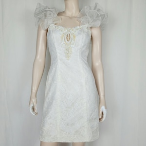 May include: A white, knee-length dress with a floral lace pattern. The dress features a sweetheart neckline, embellished with beads, and ruffled sheer sleeves. The dress is displayed on a mannequin.