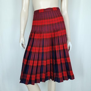 May include: A knee-length pleated skirt in a red, navy, and burgundy tartan pattern. The skirt has a classic design with evenly spaced pleats. The fabric has a slight sheen. The skirt is displayed on a mannequin. The skirt's design is reminiscent of traditional Scottish kilts.
