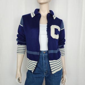 May include: A vintage navy blue zip-up sweater with a white "C" patch on the chest. The sweater features white and light blue stripes on the cuffs, collar, and hem. The sweater is displayed on a mannequin.