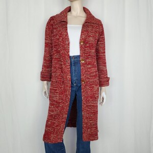 May include: A long, red and beige knitted coat with a collared neckline and three large brown buttons. The coat has long sleeves with folded cuffs and two front pockets. The mannequin is wearing blue jeans and a white top.