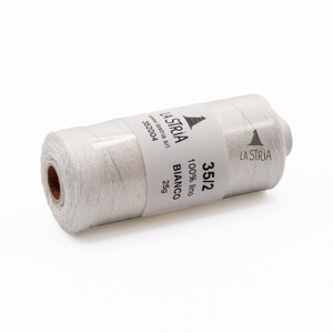 Può includere: Una bobina di filo bianco con un'etichetta che recita "LA STRIA" e "35/2 100% lino BIANCO 25g". Il filo è avvolto in plastica trasparente.