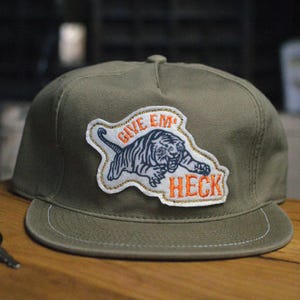 May include: Olive green flat-brimmed baseball cap with a patch featuring a tiger illustration and the words "GIVE EM HECK" in orange. The cap has a structured crown and a flat brim.