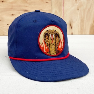 Peut inclure: Une casquette de baseball bleu marine avec une bordure en corde rouge et un patch rond représentant un cobra jaune avec des flammes rouges sur fond blanc.