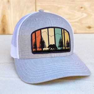 May include: A gray and white trucker hat with a patch featuring a retro-style mountain scene with a bear and trees. The patch has a colorful sunset background with orange, yellow, green, and blue hues.