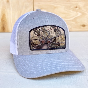 May include: A gray and white trucker hat with a black patch featuring an illustration of a giant squid attacking a ship. The patch is sewn onto the front of the hat.