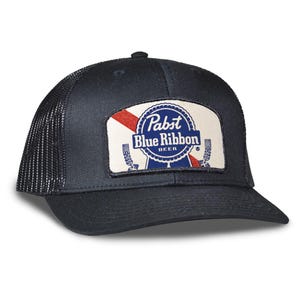 May include: A navy blue trucker hat with a mesh back and a curved brim. The front features a rectangular patch with the Pabst Blue Ribbon Beer logo in blue, red, and white. The hat has a snapback closure.