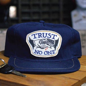 Peut inclure: Casquette de baseball bleu marine avec un écusson portant l'inscription "TRUST NO ONE". L'écusson représente une poignée de main avec un serpent et une goutte de sang. La casquette a une visière plate et une couture dorée.
