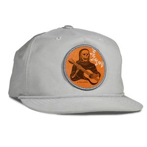 May include: A light gray baseball cap with a round patch featuring a skeleton playing a guitar. The patch is orange with brown and white details, and the text "A CALAVERA" is at the bottom. The cap has a curved brim and a rope detail.