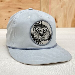 May include: A light gray baseball cap with a round, gray patch featuring a black and white illustration of an owl. The patch has a white background and a gray border.