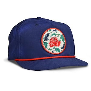 May include: A navy blue baseball cap with a red brim and a circular patch. The patch features a red flower with green leaves and blue accents on a cream background. The cap is a classic style.