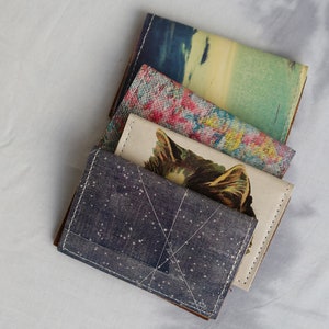 May include: Four different patterned fabric wallets stacked on top of each other. The top wallet has a blue and white starry pattern, the second wallet has a white background with a black and brown cat illustration, the third wallet has a pink and yellow floral pattern, and the bottom wallet has a blue and green watercolor pattern.