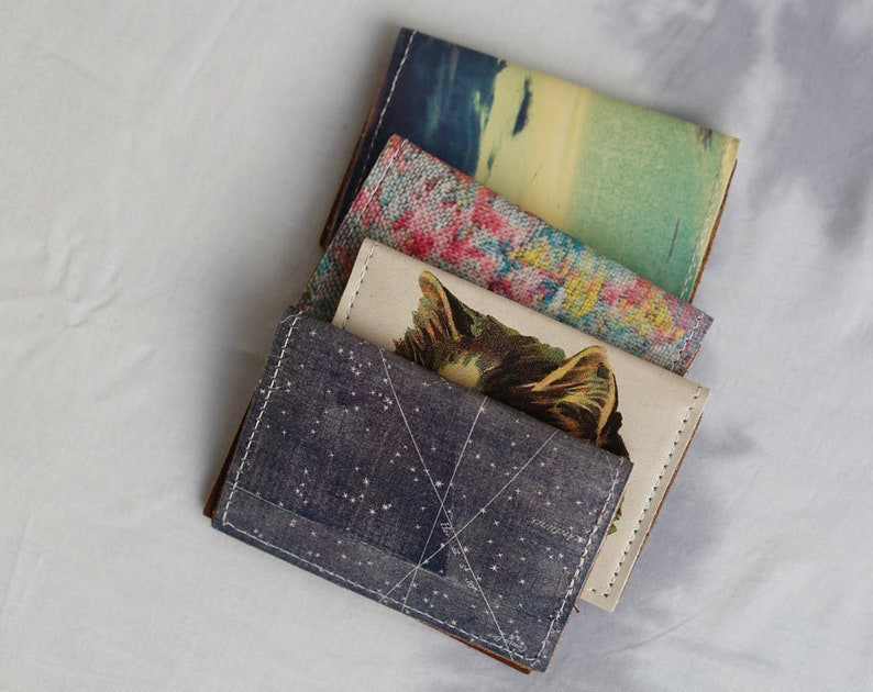 May include: Four different patterned fabric wallets stacked on top of each other. The top wallet has a blue and white starry pattern, the second wallet has a white background with a black and brown cat illustration, the third wallet has a pink and yellow floral pattern, and the bottom wallet has a blue and green watercolor pattern.