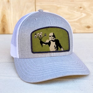 May include: A gray and white trucker hat with a green patch featuring a black and white illustration of a Stormtrooper holding a bouquet of white flowers.