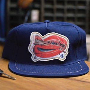 May include: A navy blue baseball cap with a flat brim. The cap features a patch with a red and white airplane design, outlined with gold stitching. The cap has a classic six-panel design.