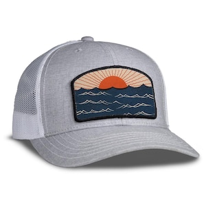 May include: Gray trucker hat with a white mesh back and a curved brim. The front features a rectangular patch with a sunset over the ocean design in orange, blue, and beige. The hat has a snapback closure.
