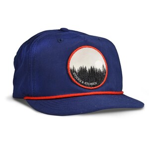 May include: A navy blue baseball cap with a red rope detail and a curved brim. The cap features a circular patch with a forest scene and the text "STICKS & STONES". The hat is a classic style.