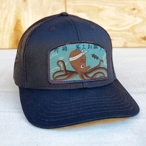 May include: Black trucker hat with a blue and brown embroidered patch featuring an octopus with a headband and holding a skewer of food. The patch has Japanese text that reads "海  え  お  魚".
