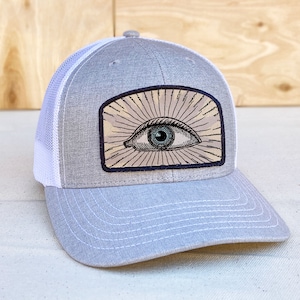 May include: A grey and white trucker hat with a patch featuring a black and white illustration of an eye with sun rays radiating from it.