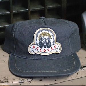 May include: A dark grey baseball cap with a patch depicting a detailed illustration of a figure wearing a crown of thorns, with the word "BELOVED" on a banner. The cap has a curved peak and gold stitching.
