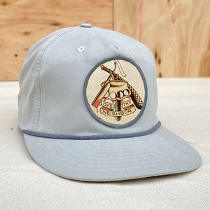 May include: A light blue baseball cap with a white patch featuring a tattoo-style design of a knife, knuckles, and the text "Love Thy Neighbour".