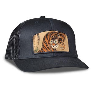 May include: Black trucker hat with a mesh back and a curved brim. The front features a rectangular patch with a detailed illustration of a tiger in orange and black stripes. The tiger is depicted in a crouching pose.