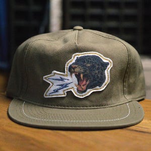 May include: Olive green baseball cap with a flat peak. The cap has an embroidered patch of a snarling black panther with blue lightning bolts. The patch is outlined with gold stitching.