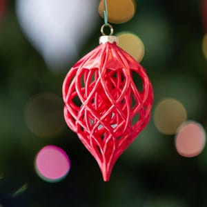 May include: A vibrant red, lattice-design Christmas ornament hangs against a blurred background of green and colorful bokeh lights. The ornament has a teardrop shape with intricate, openwork detailing, suspended by a small gold-colored cap.