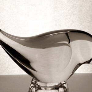 May include: A clear glass sculpture of a bird with a long, curved beak. The sculpture is perched on a clear glass base.